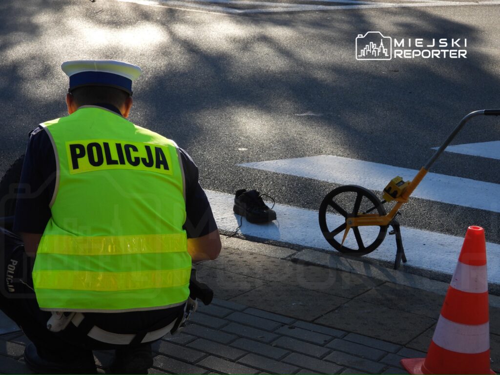 Policjant w kamizelce odblaskowej zabezpiecza miejsce zdarzenia na przejściu dla pieszych, obok stożka i koła pomiarowego.
