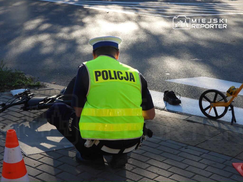 Policjant w odblaskowej kamizelce dokumentuje ślady na jezdni, obok leżą pachołki i sprzęt pomiarowy.