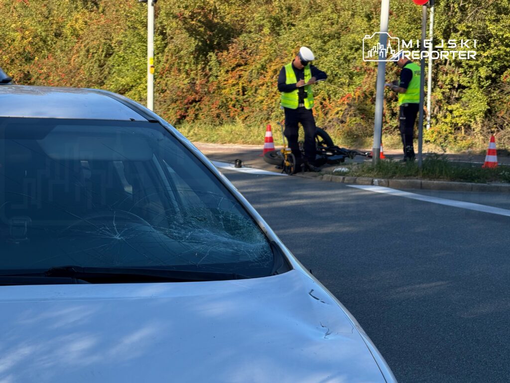 Samochód z pękniętą przednią szybą stoi przy drodze, a dwóch pracowników w kamizelkach odblaskowych zabezpiecza miejsce