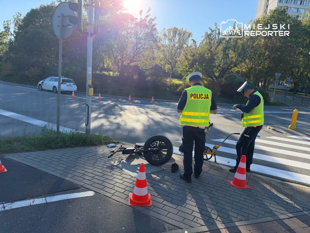 Dwóch policjantów w kamizelkach odblaskowych zabezpiecza przewrócony skuter przy przejściu dla pieszych, obok stoją pachołki.