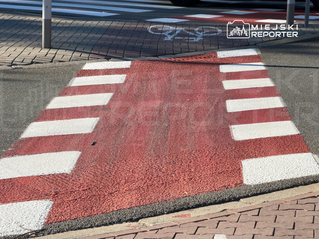 Czerwony przejazd rowerowy z białymi pasami między chodnikami, widoczny przy krawężniku i barierkach.