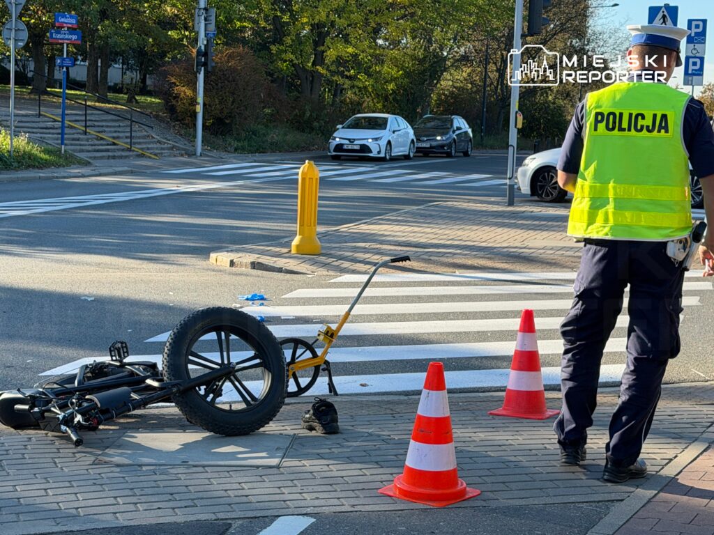 Policjant zabezpiecza miejsce zdarzenia przy przewróconym rowerze, oznaczone pachołkami, obok przejścia dla pieszych.