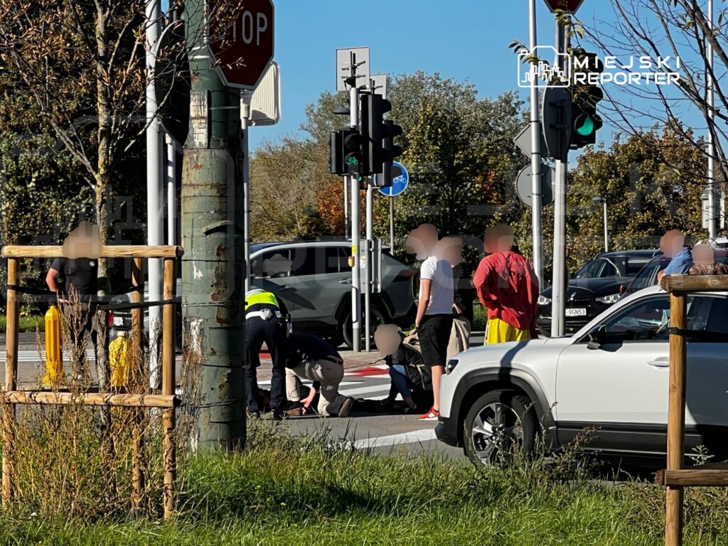 Policjant udziela pomocy osobie leżącej na przejściu dla pieszych, obok stoją piesi i samochody.