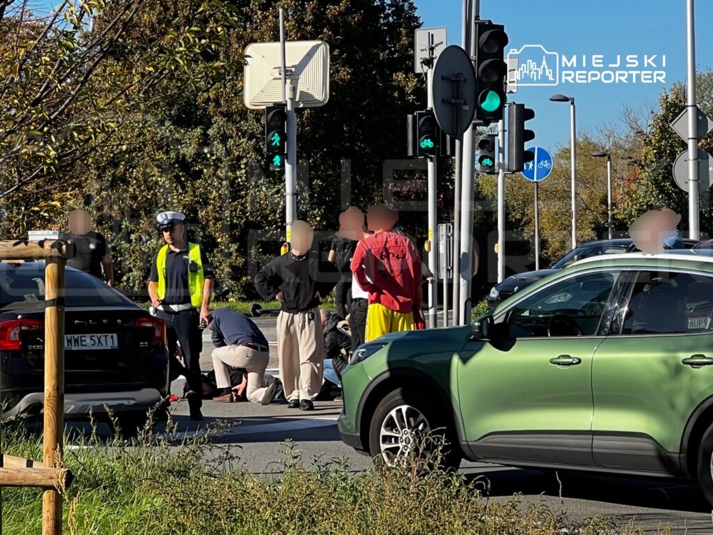 Policjanci i osoby postronne przy zatrzymanych samochodach na skrzyżowaniu, obok przejścia dla pieszych i sygnalizatorów.