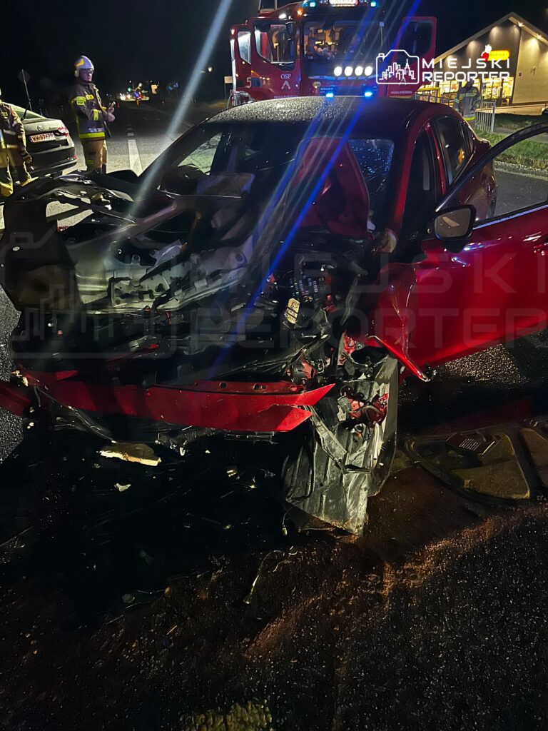 Rozbity czerwony samochód z mocno uszkodzonym przodem na nocnej drodze, obok stoją strażak i wóz strażacki.