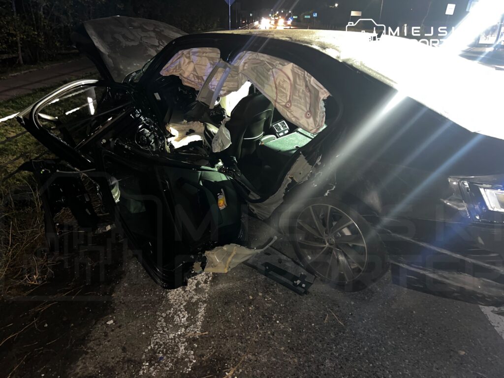 Rozbity czarny samochód z otwartymi drzwiami i wystrzelonymi poduszkami powietrznymi na nocnej drodze