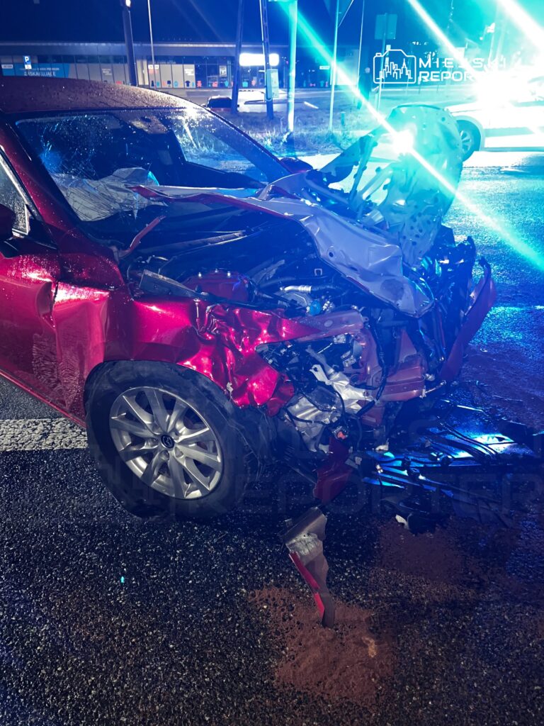 Czerwony samochód z rozbitą przednią częścią stoi nocą na mokrej jezdni, oświetlony niebieskimi światłami służb ratunkowych.