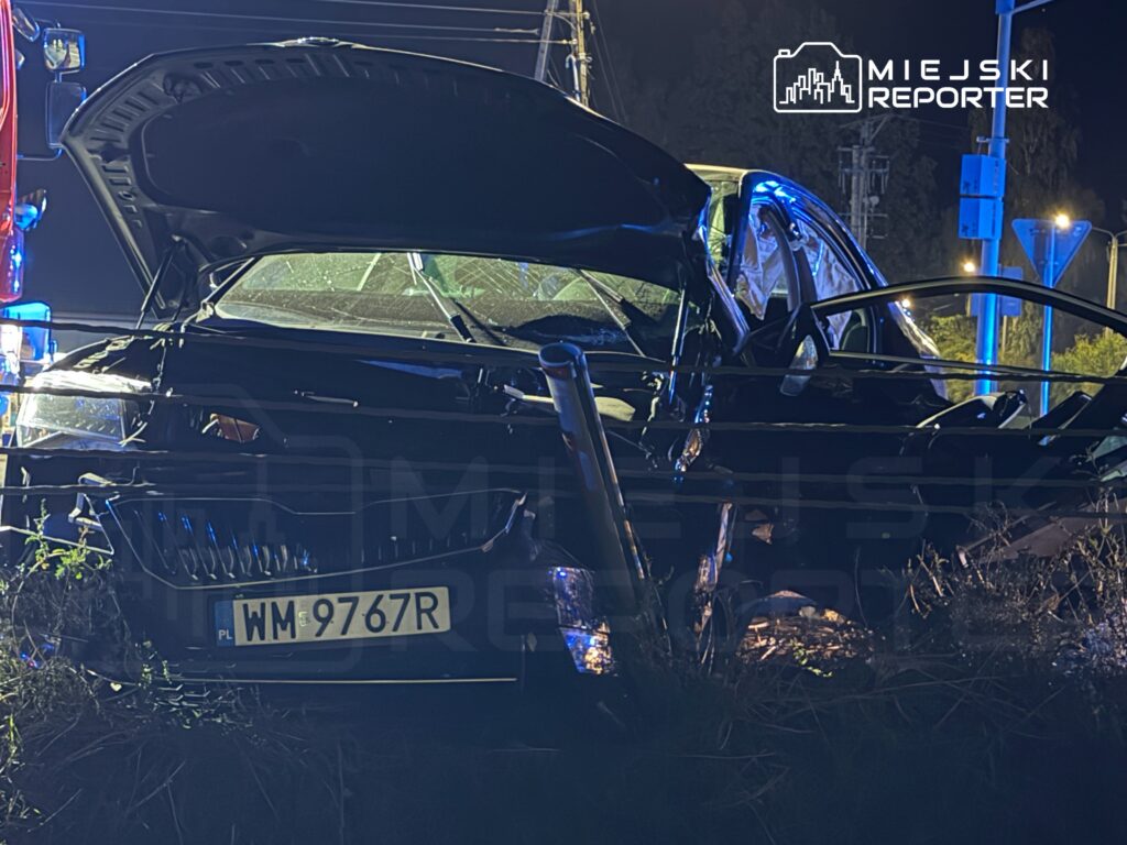 Rozbity czarny samochód z otwartą maską i drzwiami stoi w zaroślach przy oświetlonej drodze nocą.