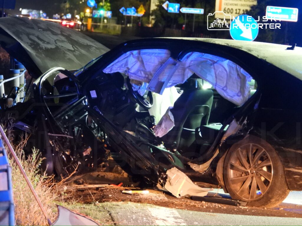 Rozbity czarny samochód z otwartą poduszką powietrzną stoi przy nocnej drodze, obok leżą odłamki karoserii.
