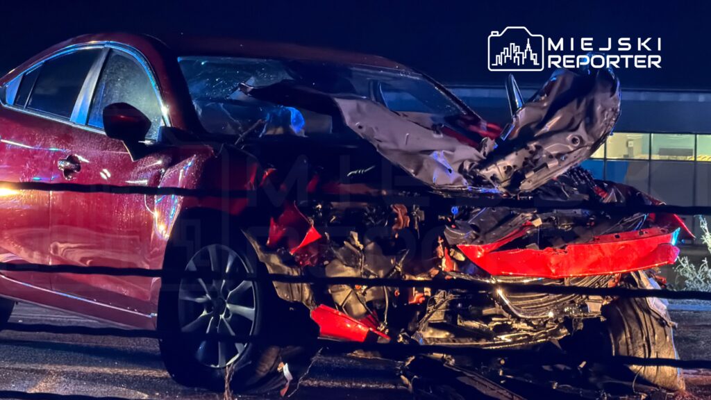 Czerwony samochód z rozbitą przednią częścią stoi nocą na parkingu przy oświetlonym budynku.