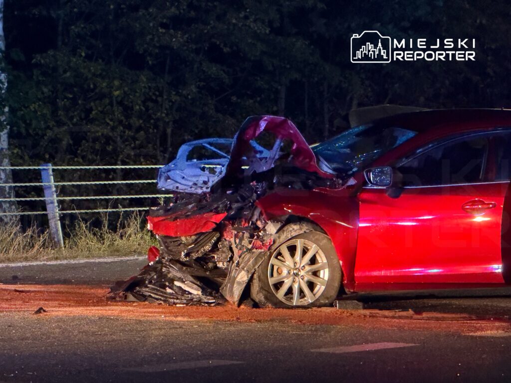 Czerwony samochód z mocno zniszczonym przodem stoi na drodze nocą, przy leśnym poboczu.