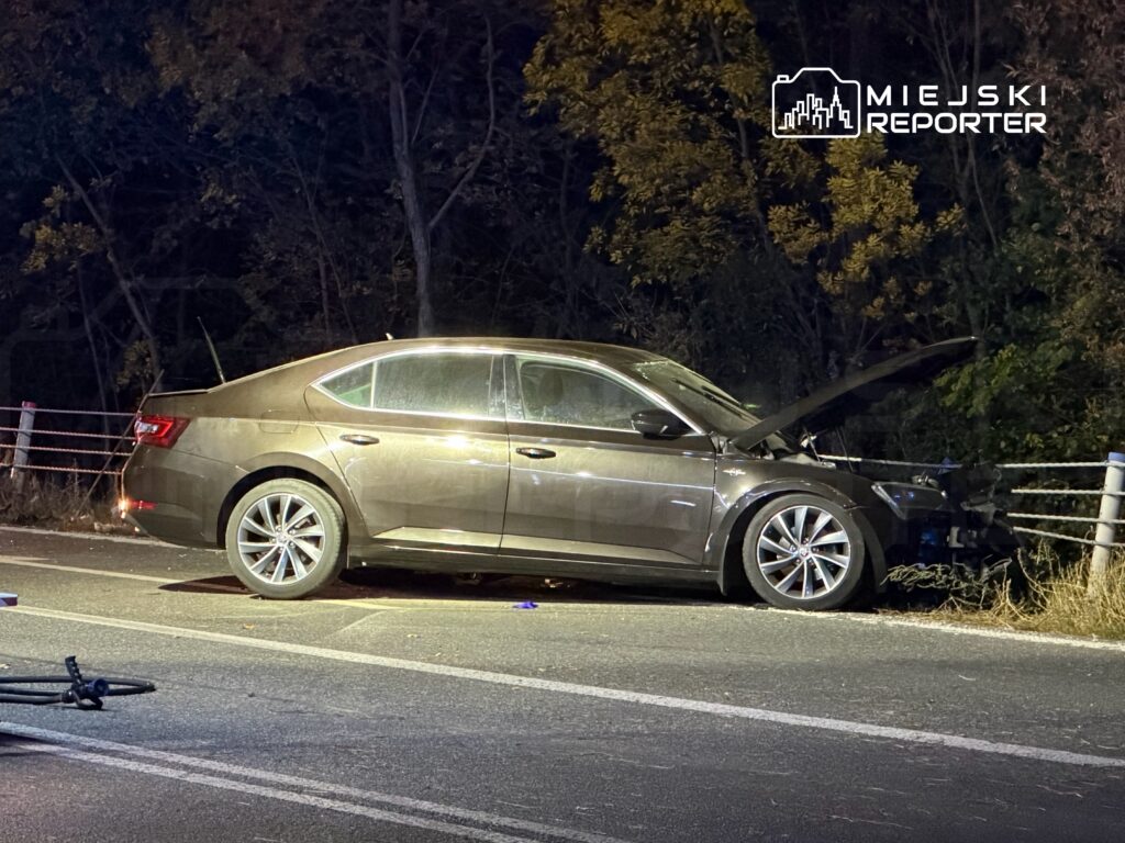 Uszkodzony samochód osobowy z otwartą maską stoi nocą przy barierce na poboczu drogi.