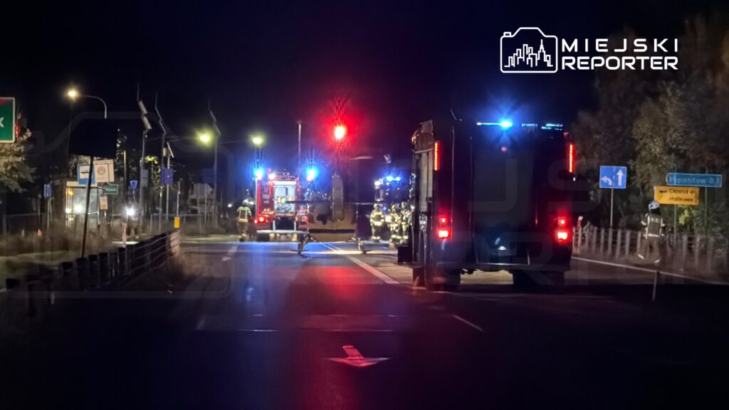 Strażacy i wozy ratunkowe na nocnej drodze, obok autobusu; skrzyżowanie zabezpieczone podczas akcji służb.