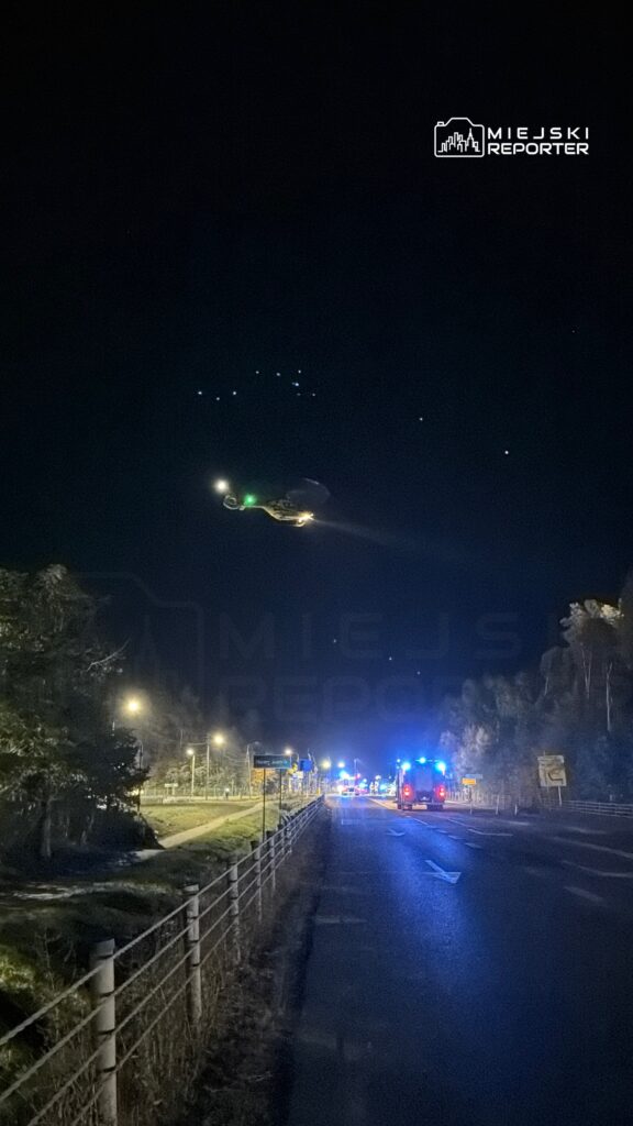 Śmigłowiec krąży nad nocną drogą, przy której stoją pojazdy z włączonymi niebieskimi światłami.