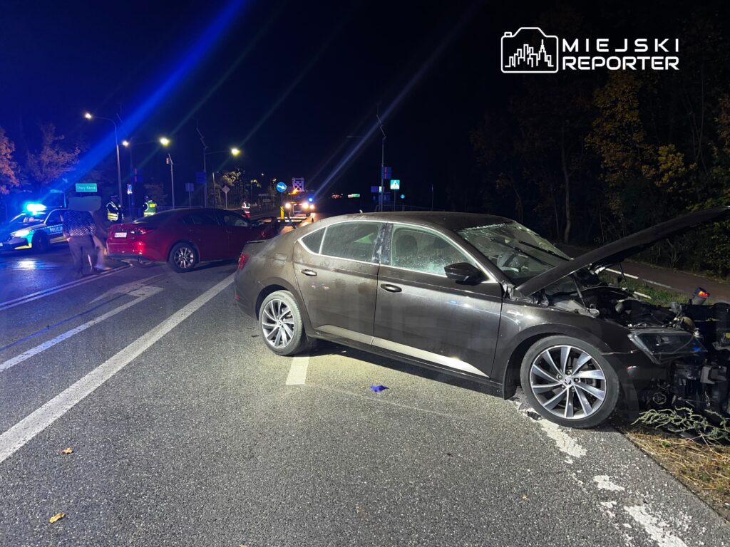 Uszkodzony samochód z otwartą maską stoi na nocnej drodze po kolizji, obok niego widać drugi pojazd i światła służb.