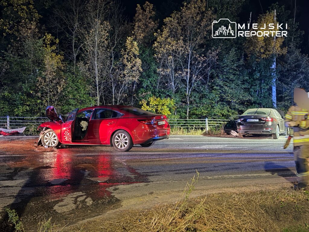 Uszkodzony czerwony samochód i szary sedan stoją na mokrej drodze, obok pracuje strażak w odzieży ochronnej.