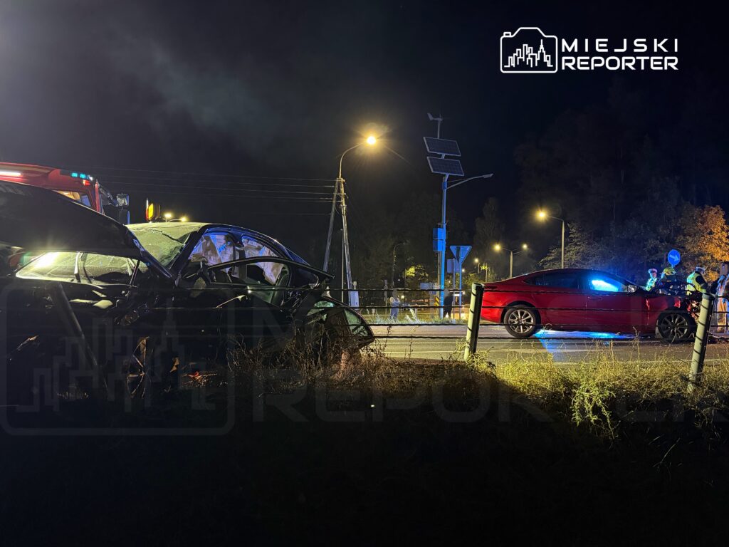 Dwa samochody po zderzeniu na nocnej drodze, obok pojazdu służb ratunkowych i oświetlonego miejsca zdarzenia.