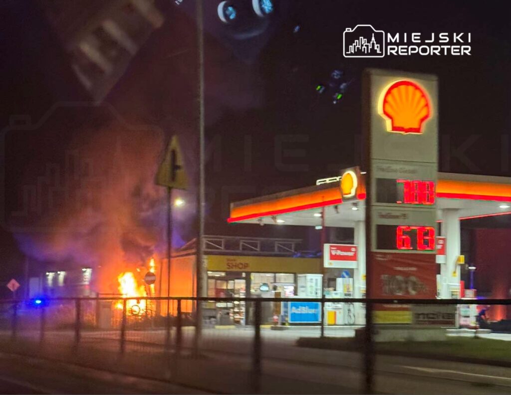 Płomienie i gęsty dym przy stacji paliw Shell, za ogrodzeniem i obok zaparkowanych pojazdów służb ratunkowych nocą.