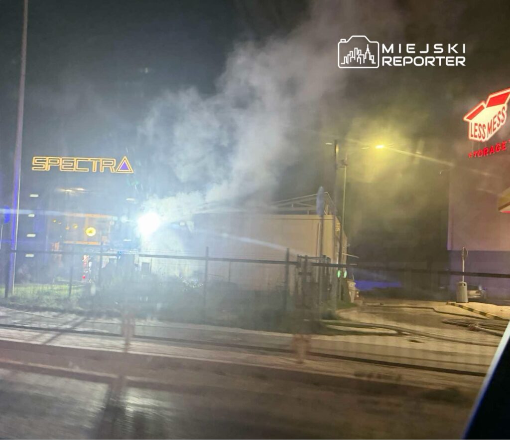 Kłęby dymu unoszą się nocą nad ogrodzonym obiektem handlowym z szyldami „SPECTRA” i „LESSNESS