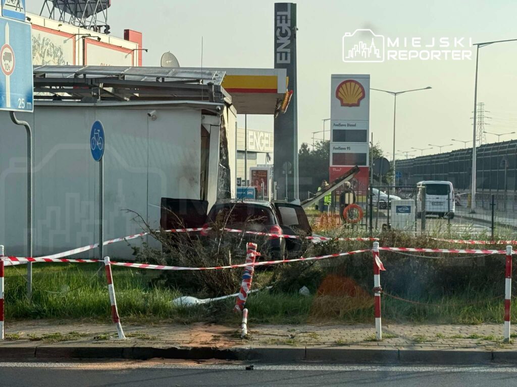 Uszkodzony samochód osobowy stoi w zaroślach za taśmą ostrzegawczą, przy stacji paliw i pawilonie handlowym.