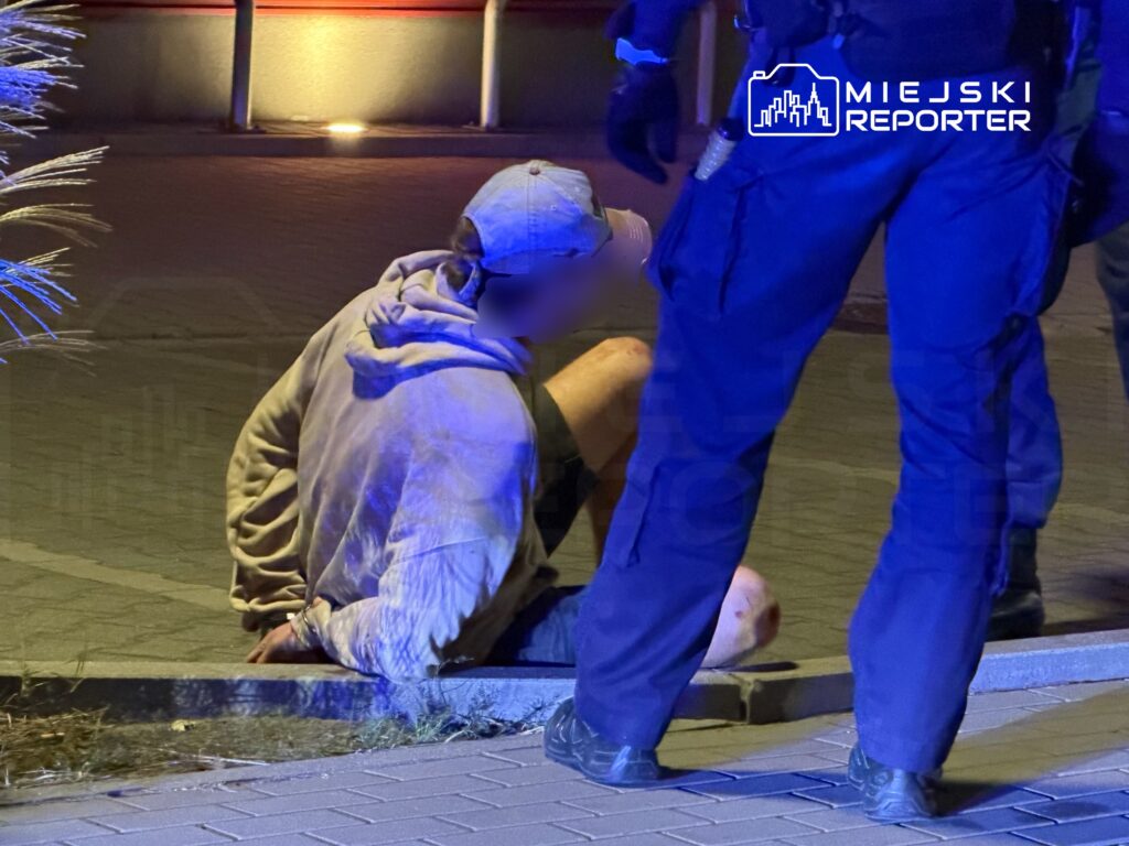 Osoba w jasnej kurtce siedzi na chodniku przy krawężniku, obok stoją funkcjonariusze w niebieskich mundurach.