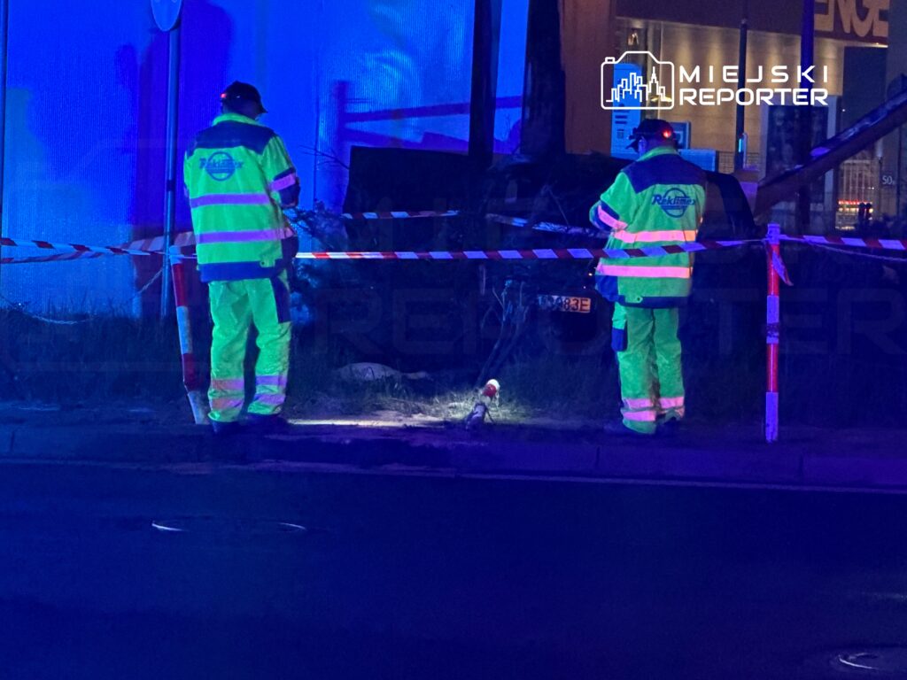 Dwaj pracownicy w odblaskowych strojach zabezpieczają taśmą teren przy chodniku, oświetlony niebieskim światłem.