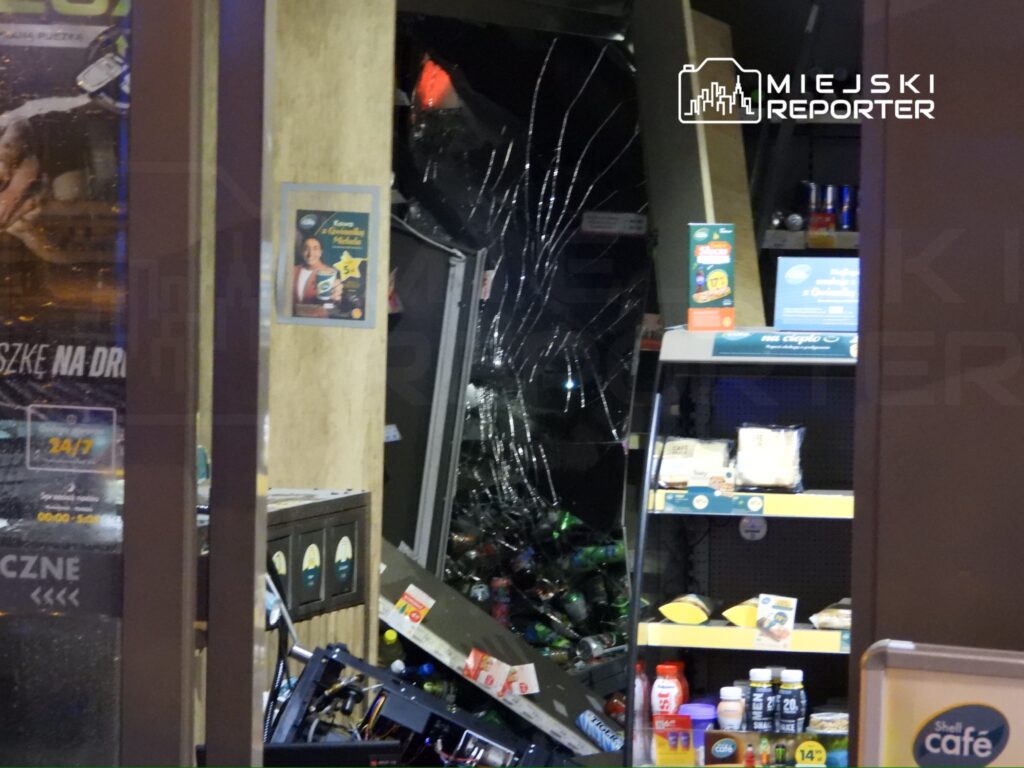 Rozbite wejście do sklepu całodobowego, z popękaną szybą i przewróconymi półkami z towarem wewnątrz.