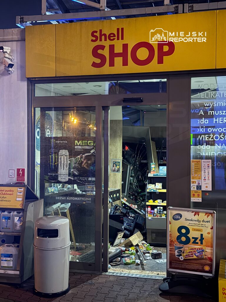 Uszkodzony sklep Shell z rozbitym wejściem i porozrzucanymi produktami na podłodze nocą.