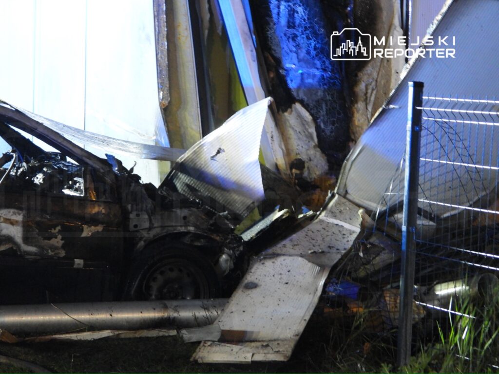 Wrak samochodu dostawczego po zderzeniu z konstrukcją przy ogrodzeniu, nocą oświetlony niebieskim światłem