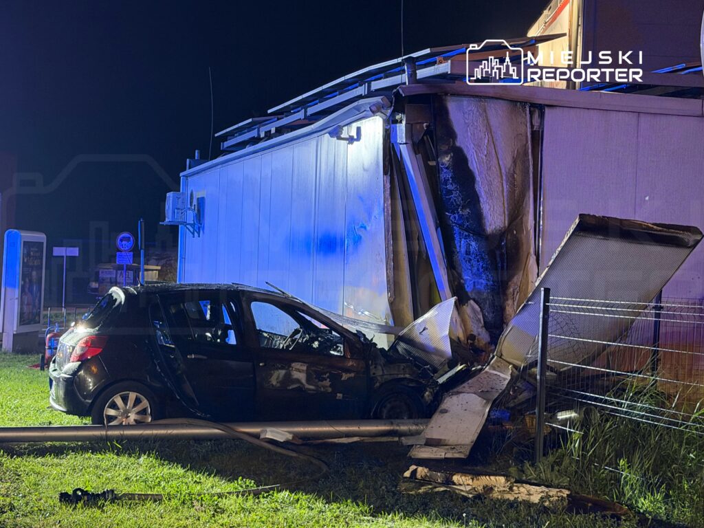 Rozbity czarny samochód przy uszkodzonej ścianie budynku, nocą, w świetle niebieskich świateł.