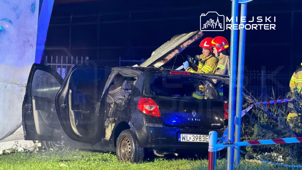 Strażacy oglądają spalonego samochód z otwartymi drzwiami podczas nocnej akcji na ogrodzonym terenie.