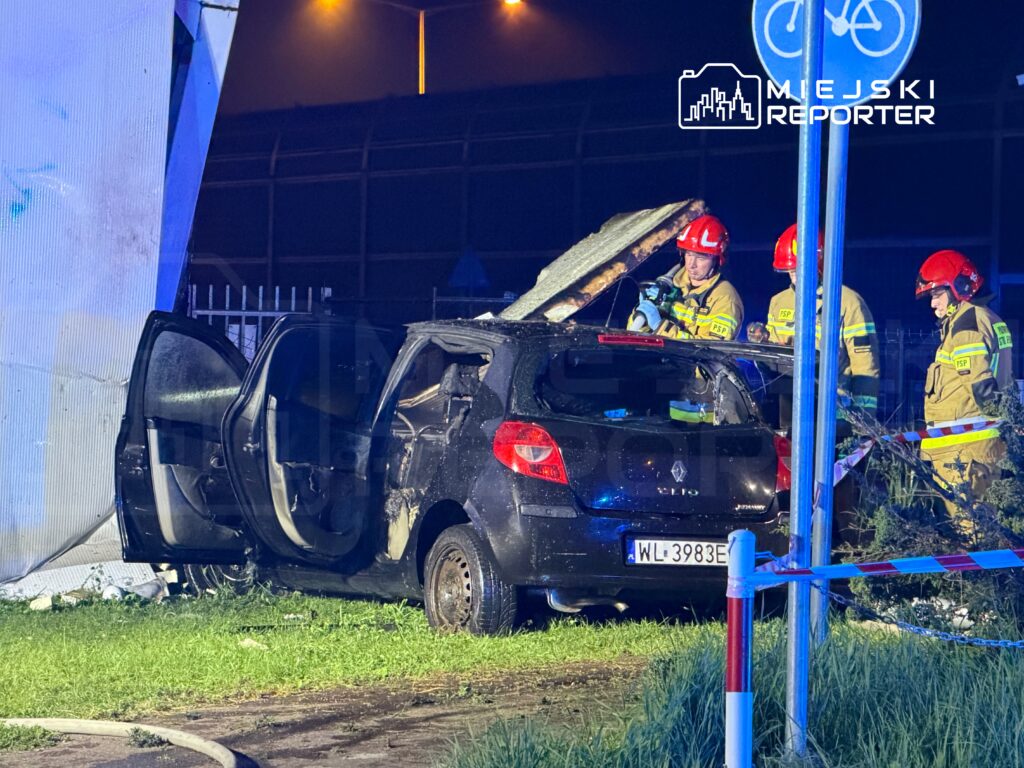 Strażacy oglądają rozbity samochód osobowy z otwartymi drzwiami przy ogrodzeniu, nocą oświetloną lampami i światłami służb.