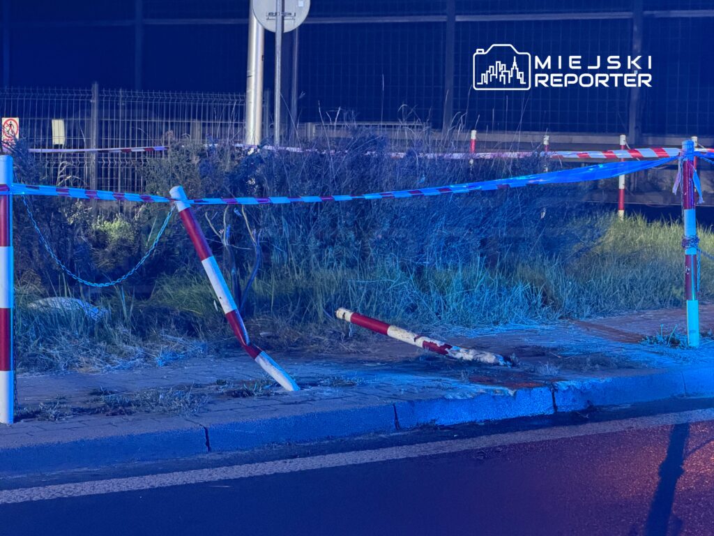 Uszkodzone czerwono-białe słupki i taśma odgradzają chodnik przy ogrodzonym, zarośniętym terenie nocą.