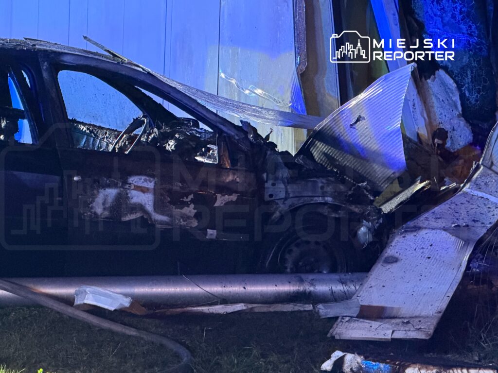 Rozbity, zniszczony samochód osobowy leży przy metalowej barierze na poboczu drogi nocą.