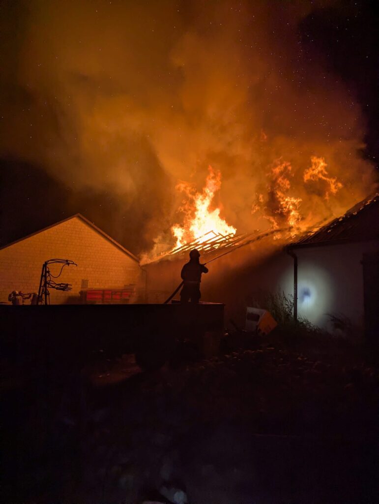 Strażak gasi płonący dach budynku mieszkalnego nocą, wśród gęstego dymu i płomieni.