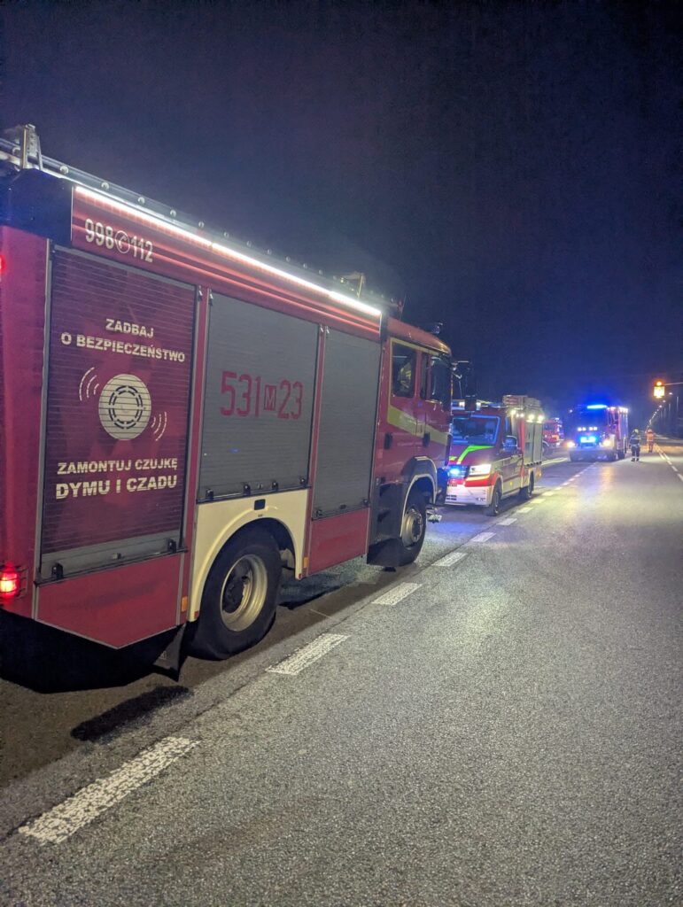 Wozy strażackie i ratunkowe stoją nocą na poboczu drogi, z włączonymi niebieskimi światłami.