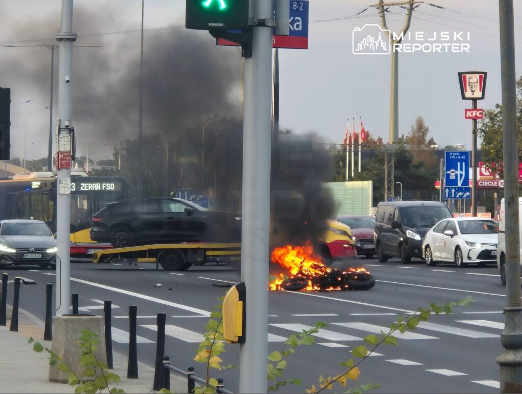 Płonący motocykl na skrzyżowaniu, z którego unosi się gęsty czarny dym; obok stoją samochody i autobus.