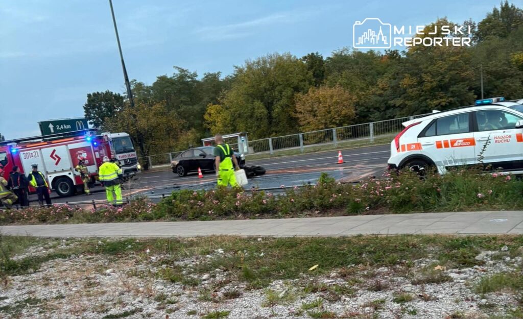 Strażacy i ratownicy zabezpieczają czarny samochód na drodze, obok wozu strażackiego i ambulansu.