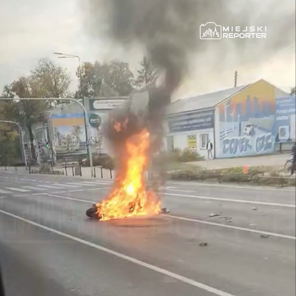 Płonący przedmiot na środku jezdni, z którego unosi się gęsty dym; w tle przystanek i budynki.