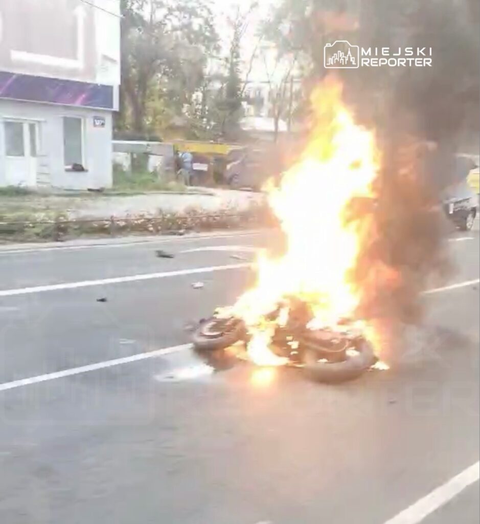 Motocykl płonie na jezdni, unoszą się nad nim płomienie i gęsty dym.
