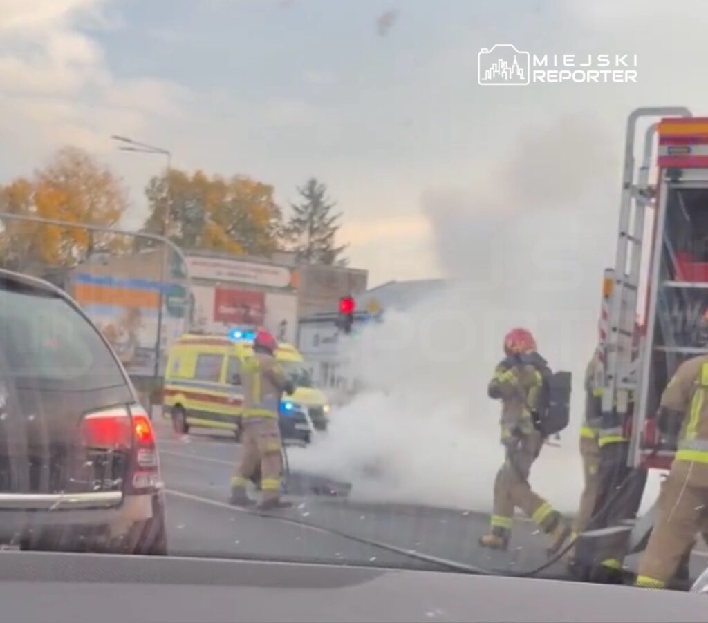 Strażacy gaszą dymiący pojazd na skrzyżowaniu, obok stoją wóz strażacki i karetka pogotowia.
