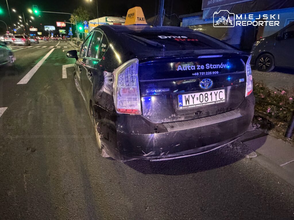 Uszkodzona z tyłu taksówka Toyota Prius stoi nocą na pasie ruchu przy skrzyżowaniu.