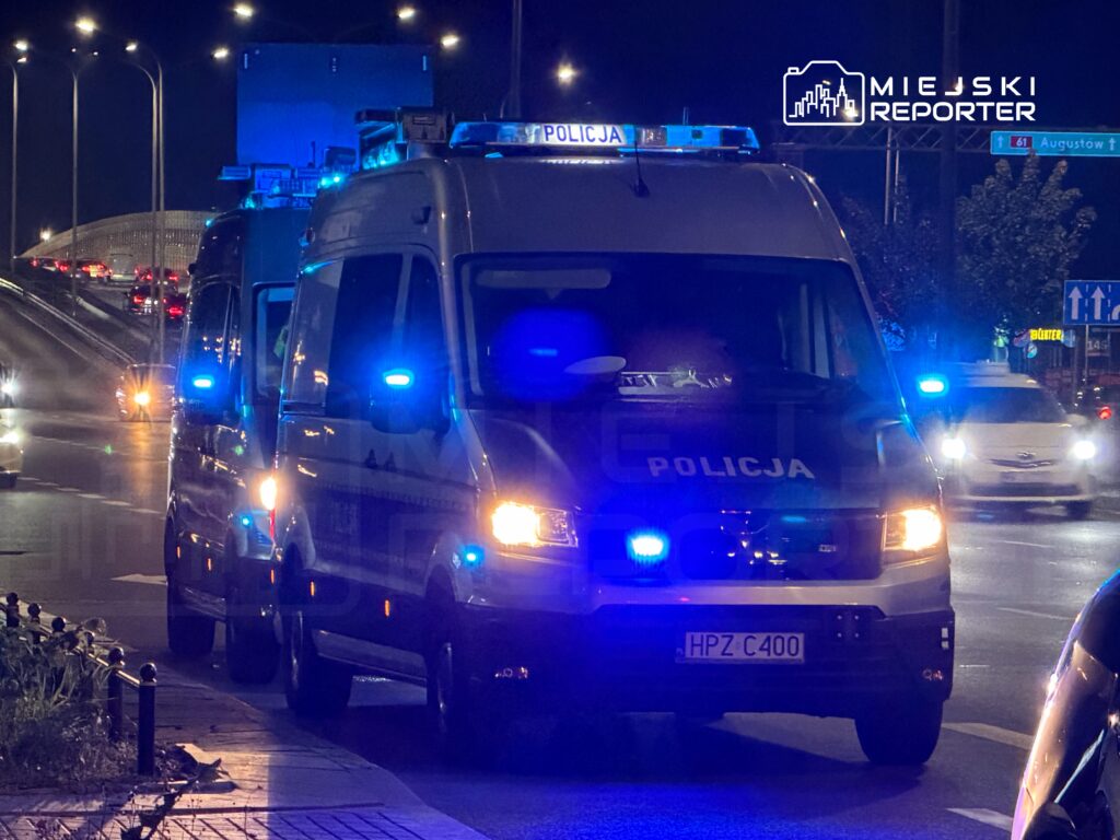 Policyjny bus z włączonymi niebieskimi światłami stoi nocą na miejskiej ulicy, obok innych pojazdów służb.