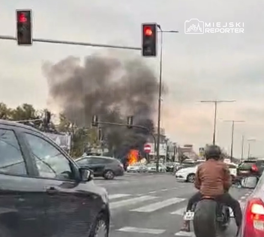 Samochód płonie na skrzyżowaniu, a nad ulicą unosi się gęsty dym; na pierwszym planie stoją auta i motocyklista.