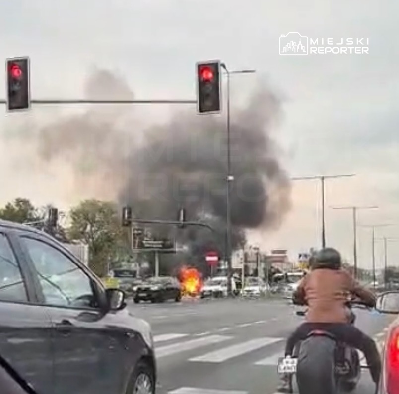 Płonący samochód na skrzyżowaniu, z którego unosi się gęsty czarny dym; na pierwszym planie motocyklista i auta.