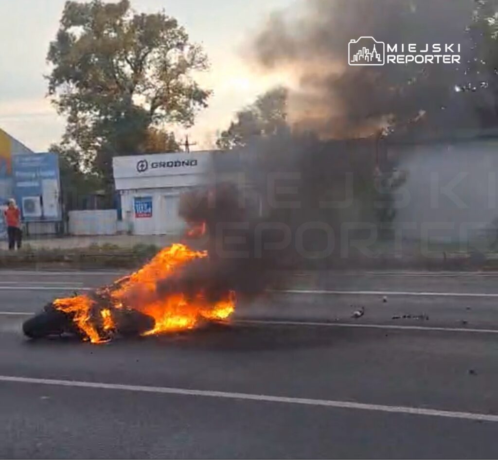 Płonący motocykl na jezdni, nad którym unosi się gęsty, ciemny dym.