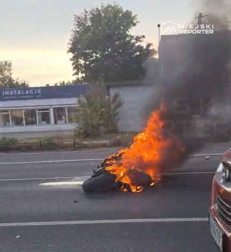Motocykl płonie na jezdni, unosząc gęsty czarny dym przy budynku usługowym.