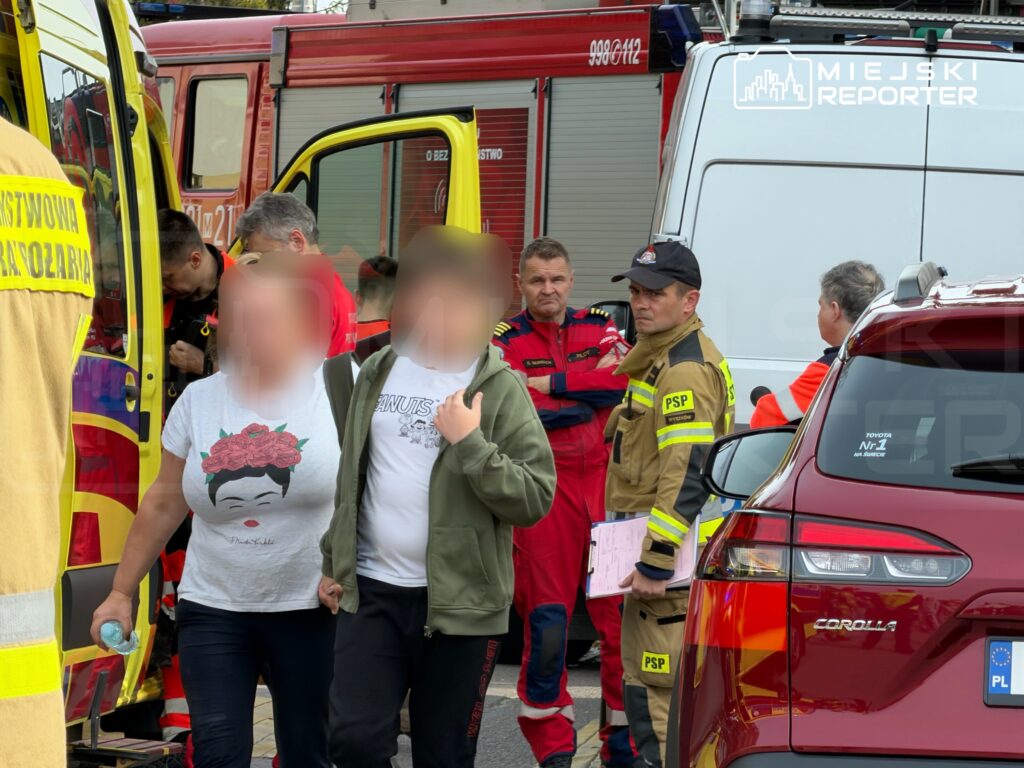 Ratownicy medyczni i strażak PSP pomagają dwóm osobom przy karetkach i samochodach ratowniczych.