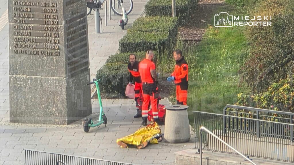 Trzech ratowników w pomarańczowych strojach stoi obok żółtego noszy na brukowanym placu przy pomniku.