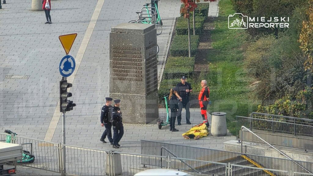 Policjanci rozmawiają z ratownikiem w odblaskowym stroju przy żółtych noszach na miejskim placu z pomnikiem i barierkami.